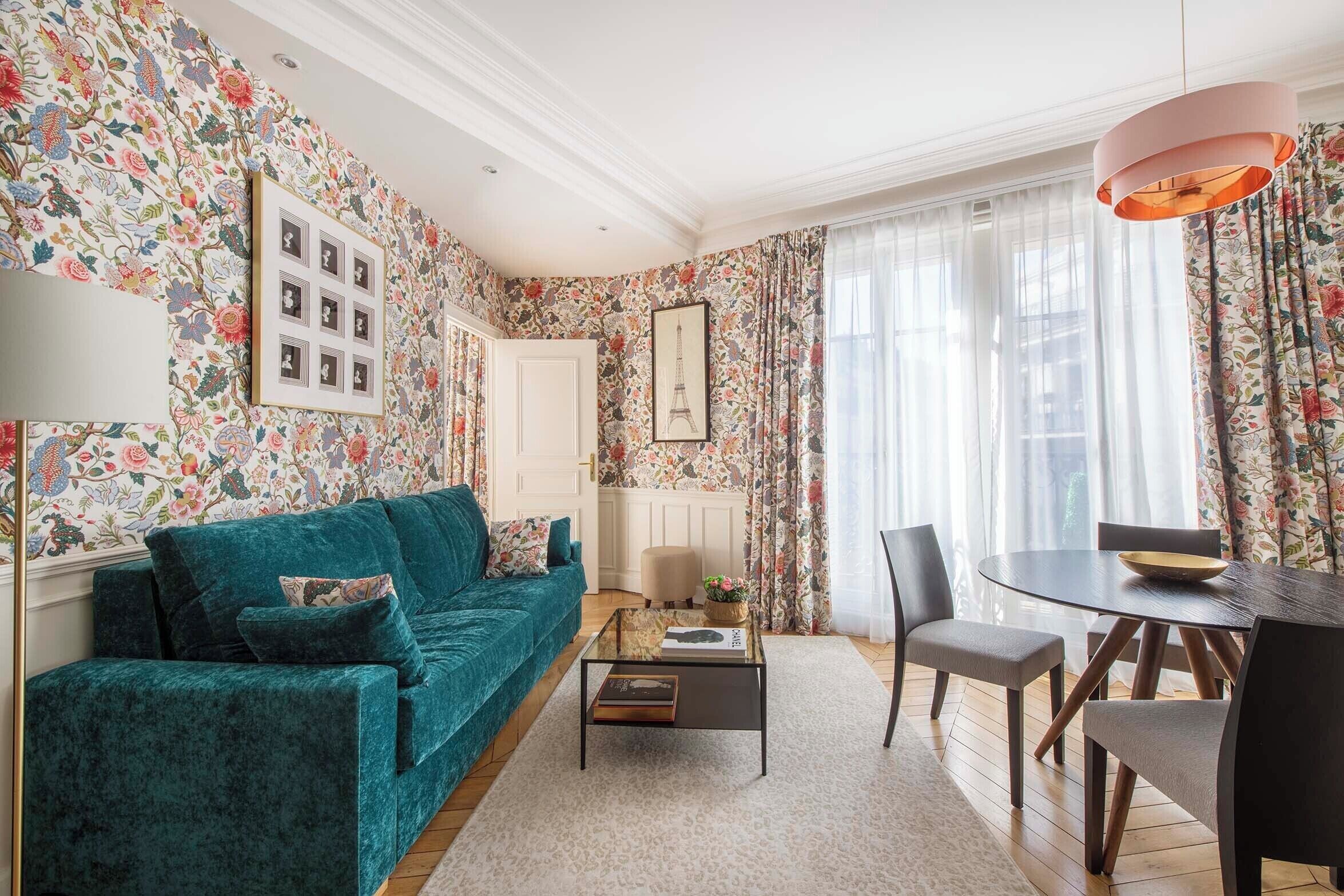 Ein Wohnzimmer mit einem salbeengrünen Sofa, einem Glastisch, einem Esstisch mit Stühlen, blumigem Wandpapier und einem großen Fenster mit Vorhängen.