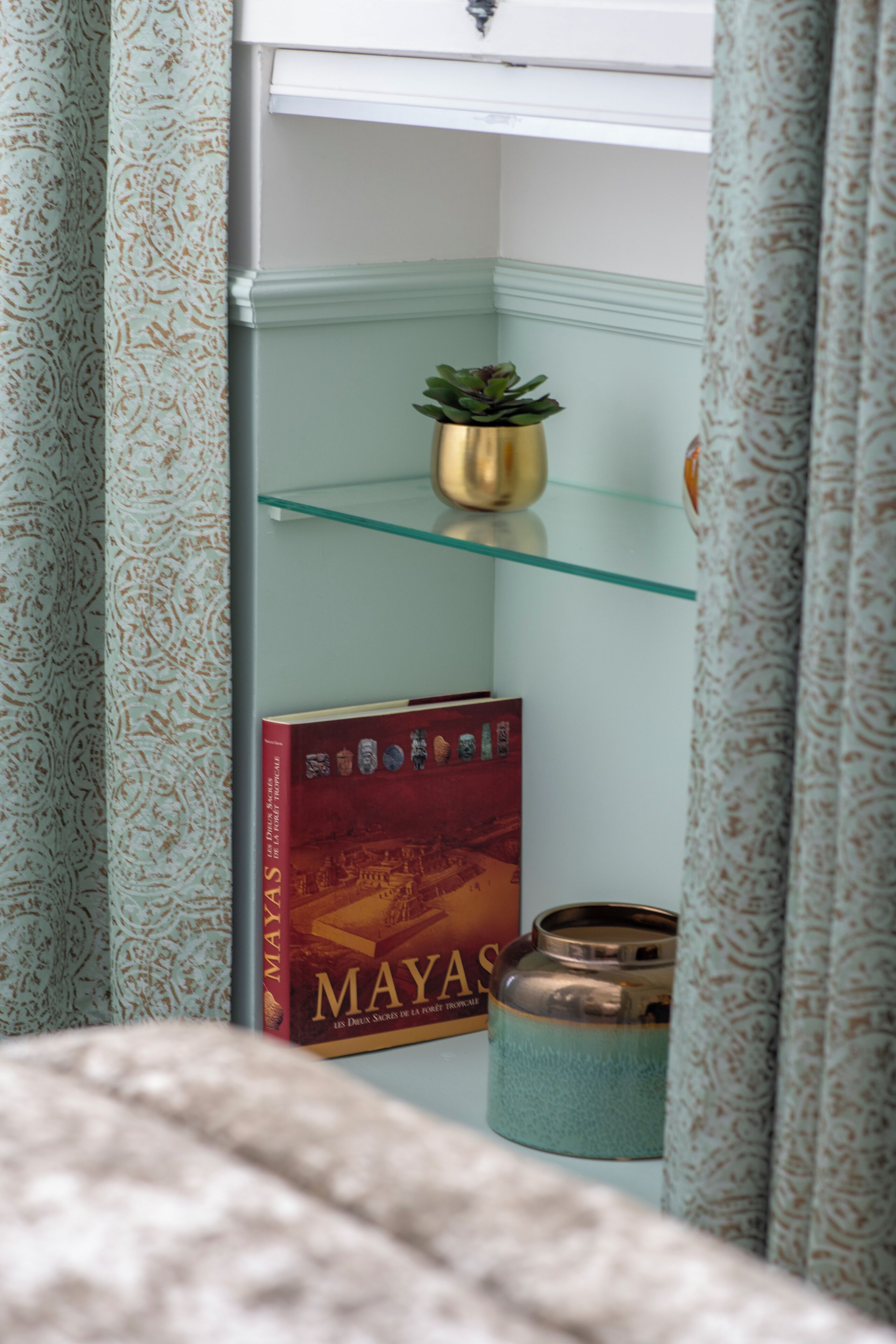Canto de um quarto com cortinas estampadas, uma prateleira de vidro com uma pequena planta, um livro intitulado 'MAYAS' e um recipiente de cerâmica.