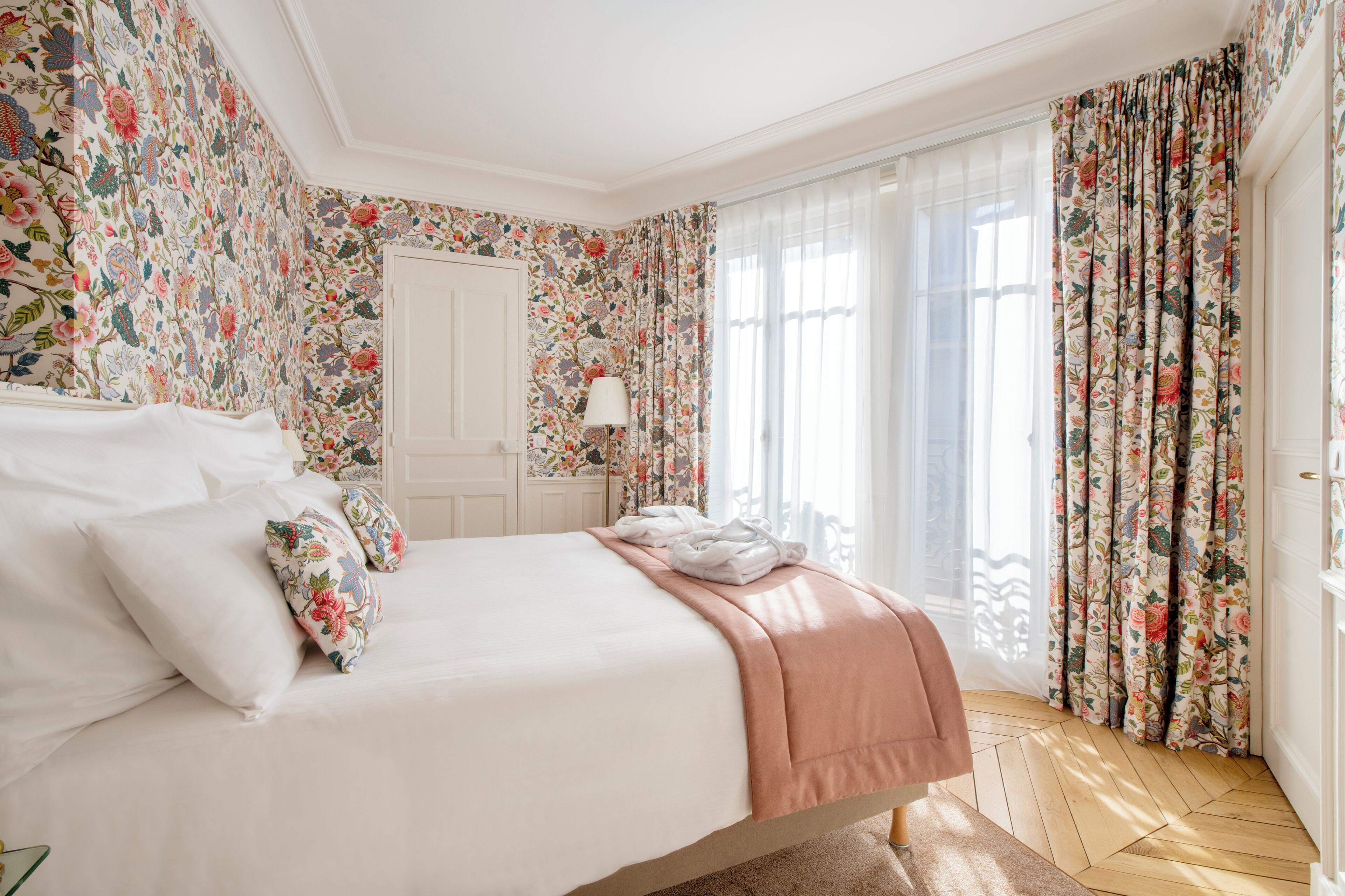 A bedroom with floral wallpaper, a bed with white and pink bedding, a nightstand, and a window with curtains.