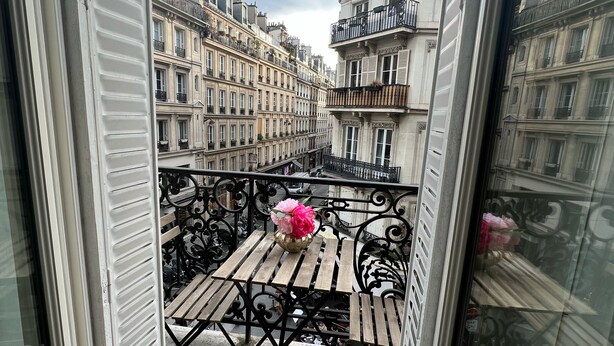 Un balcón con mesa y sillas, un florero con flores y vista a edificios parisinos.