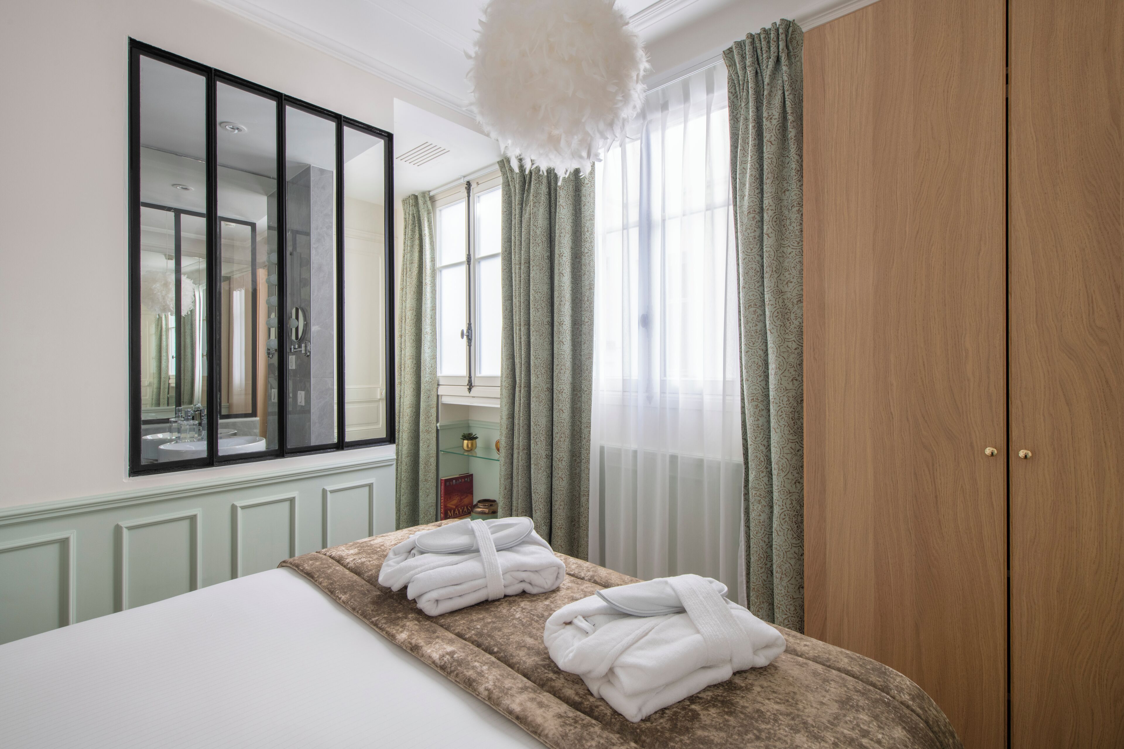 A hotel room with a large bed, a wooden wardrobe, a vanity with a mirror, and a window with curtains.