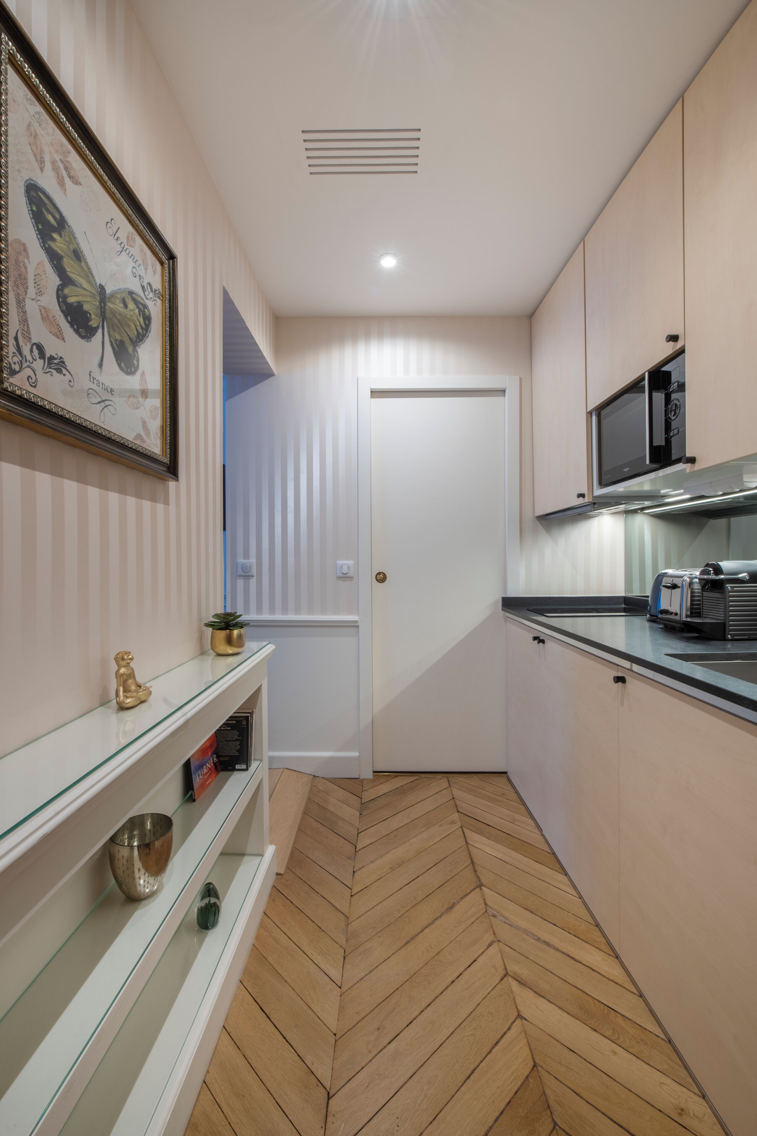 Une cuisine étroite avec un sol décoré d’un motif en chevrons, un four à micro-ondes, un grille-pain et une œuvre d’art représentant un papillon, encadrée et accrochée au mur.