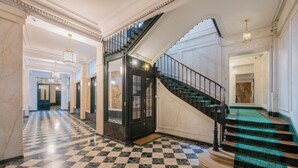 Un vestíbulo espacioso con piso de mármol a cuadros, una escalera con alfombra verde y un ascensor negro.