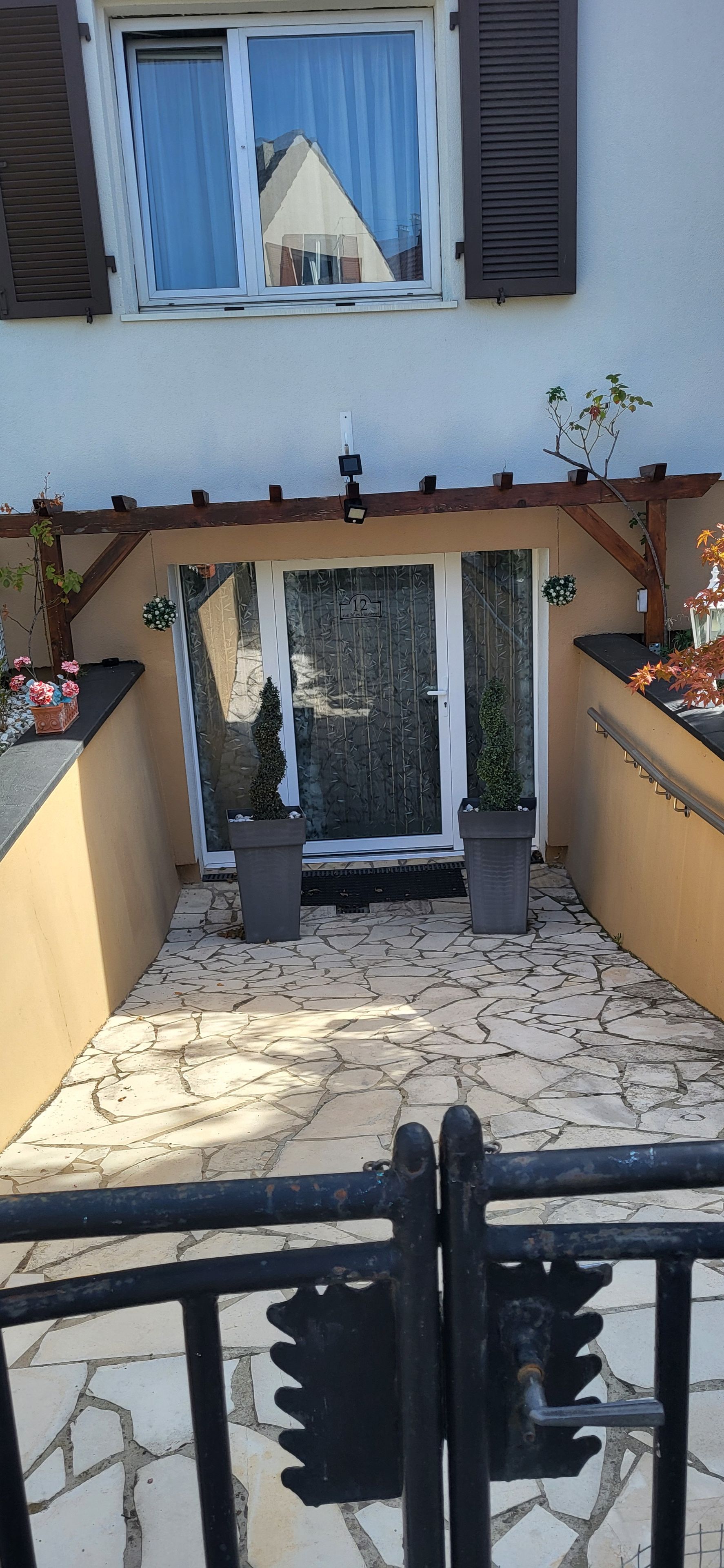 A covered patio area with a stone floor, two potted plants, a sliding glass door, and a small window with shutters.