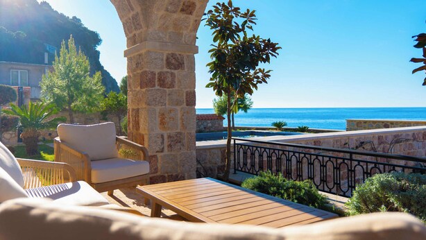 A stone archway leading to a terrace with a wooden table and chairs, overlooking a coastal view.
