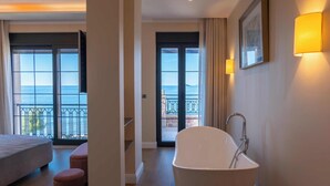 A modern bathroom with a freestanding bathtub, a large window with a view of the sea, and a balcony with a railing.