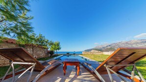A poolside area with lounge chairs and a table, overlooking a clear blue sky and a distant mountain range.