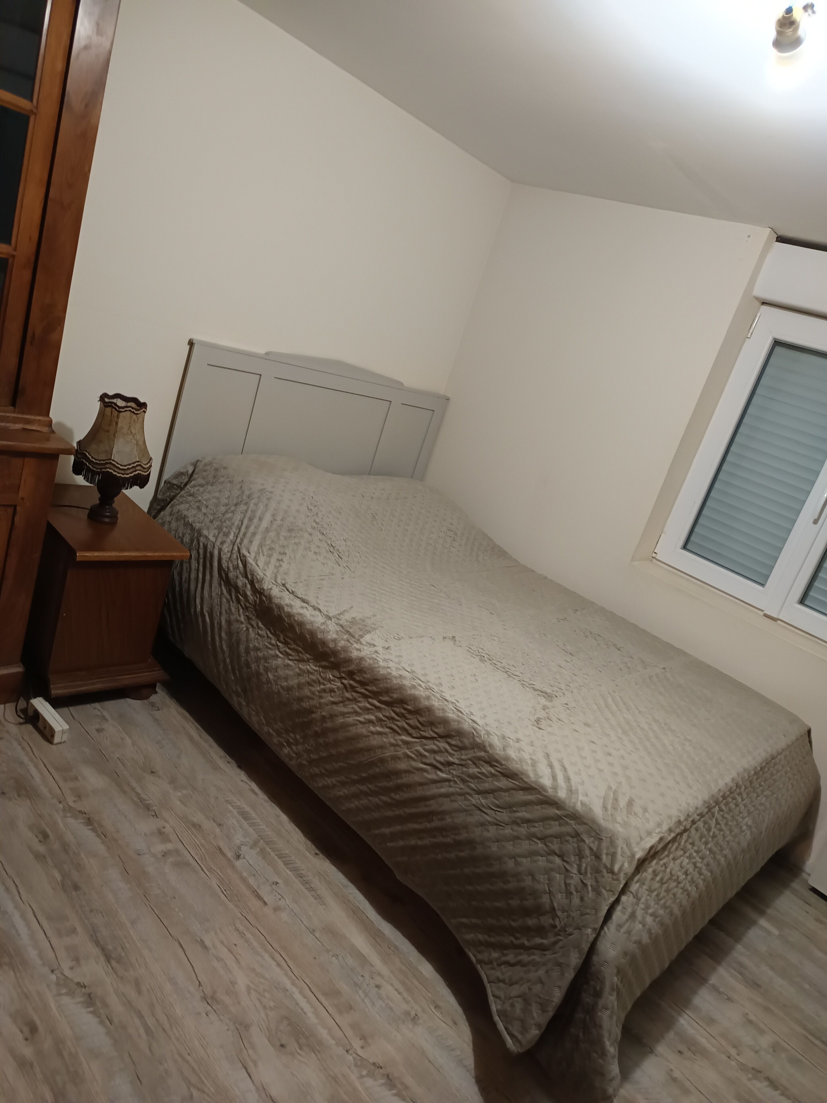 Una habitación pequeña con una cama individual, cabecera de madera, mesita de noche con lámpara y ventana con persianas.