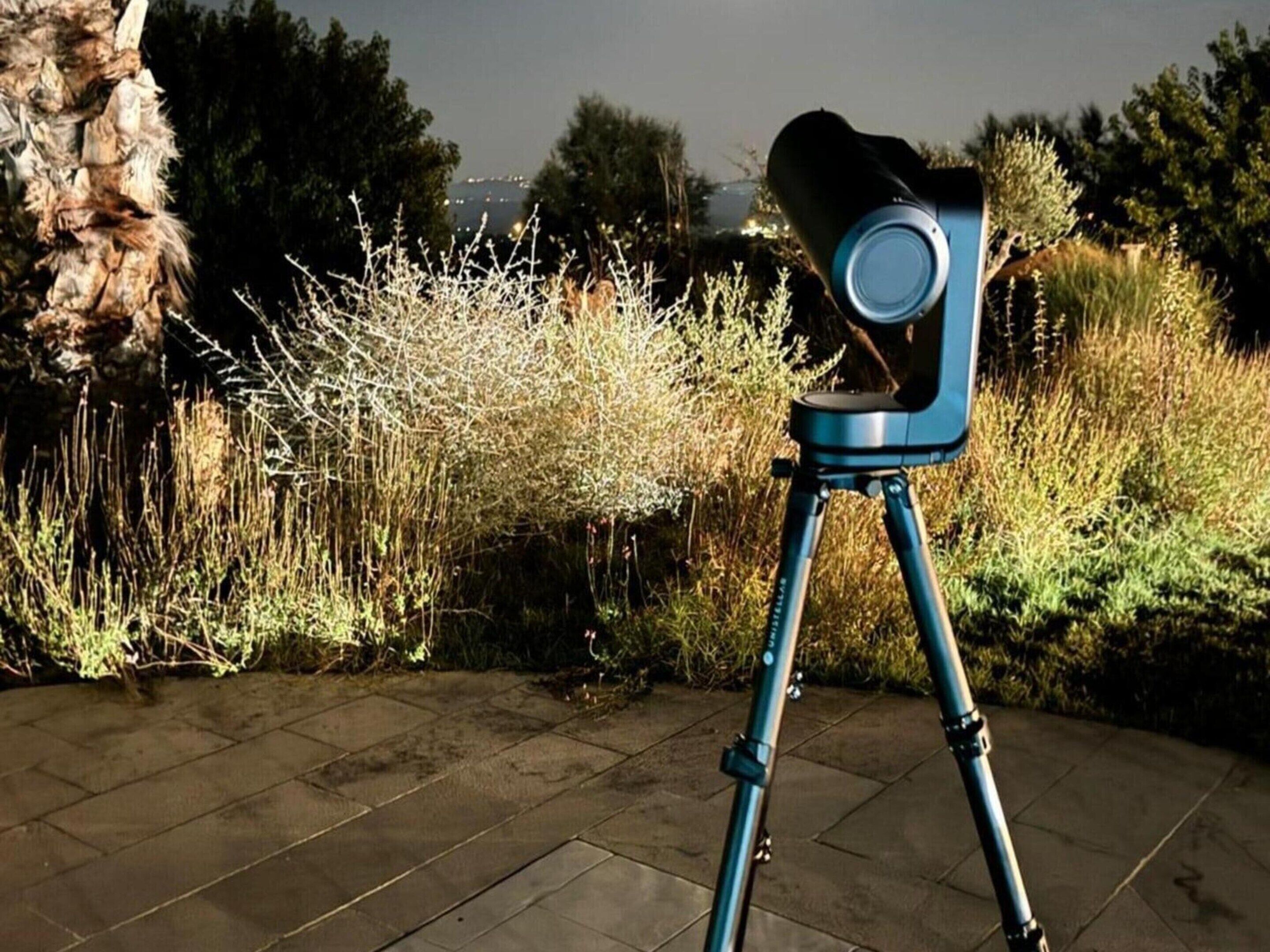 A telescope on a tripod in an outdoor setting with a stone-paved area and various plants.