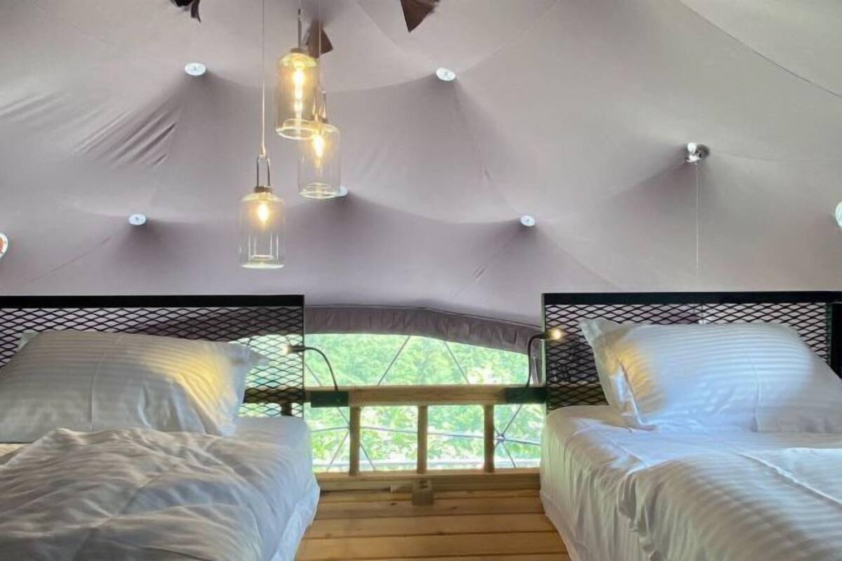 Une pièce aménagée comme une tente, avec deux lits, un luminaire suspendu et une vue sur la verdure environnante.