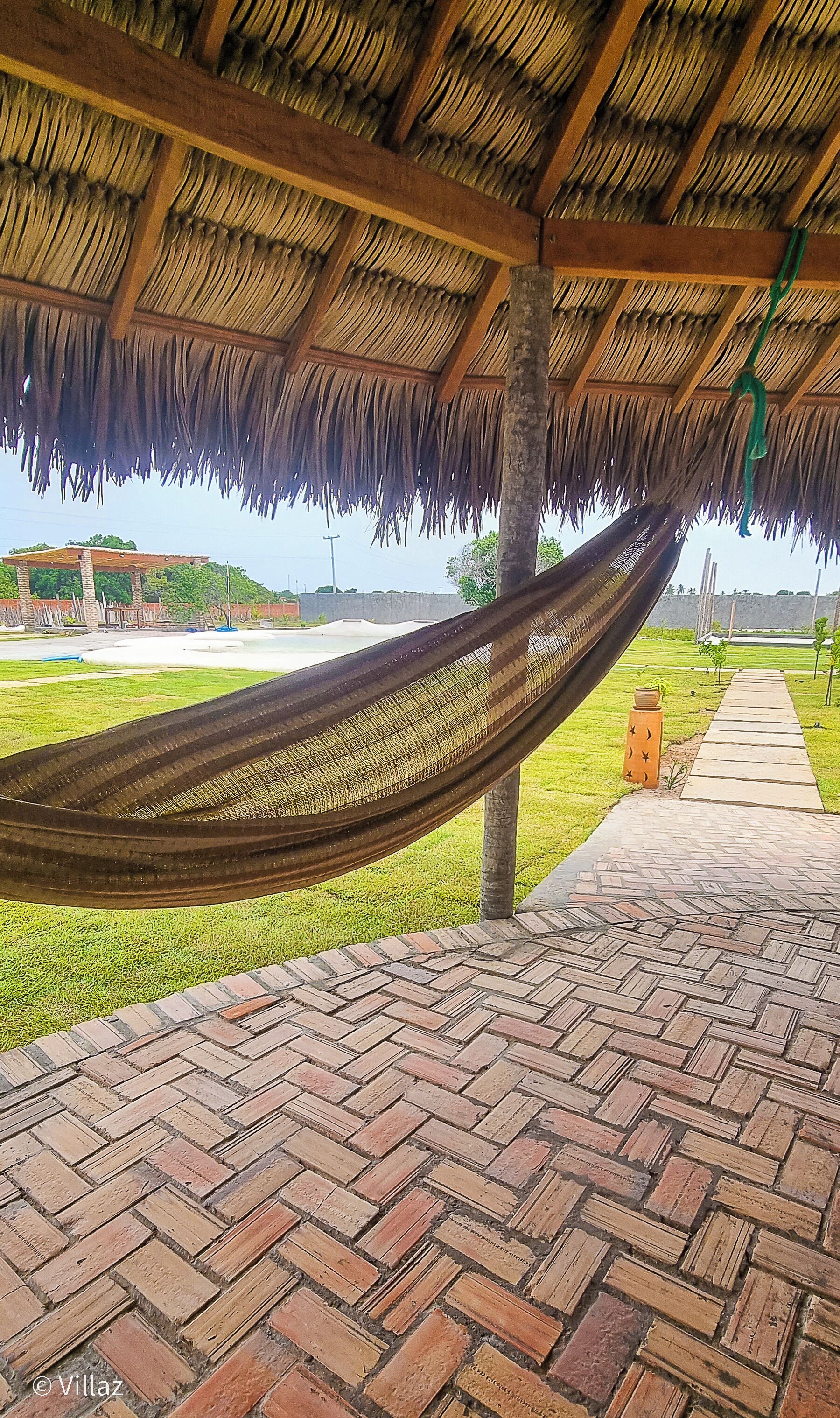 A hammock under a thatched roof, a brick pathway, and a grassy area with a view of a distant building.