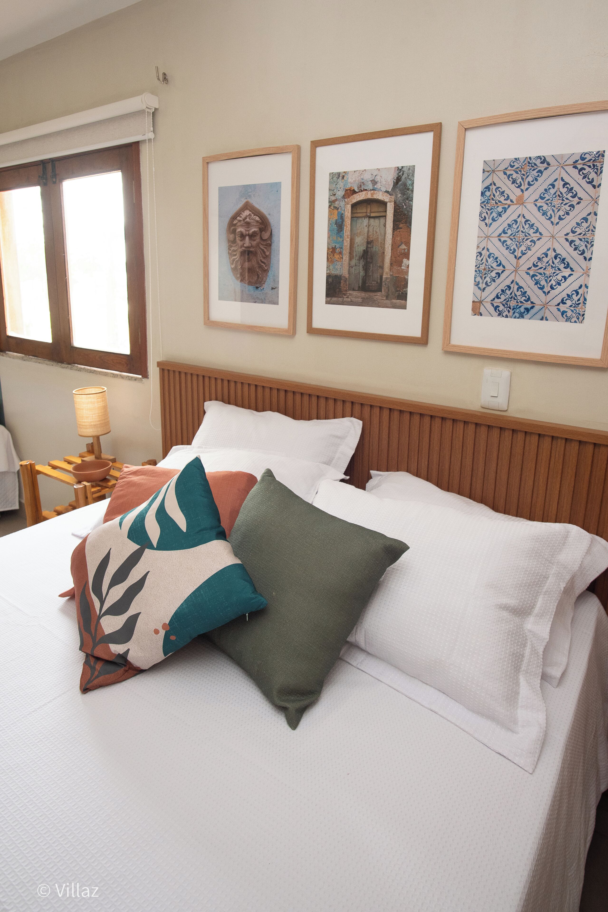 A neatly made bed with white bedding and decorative pillows, a wooden headboard, and framed artwork on the wall.