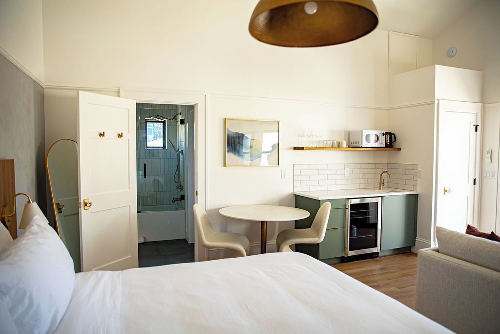 A hotel room with a bed, a small dining table with chairs, a kitchenette, and a bathroom visible through an open door.