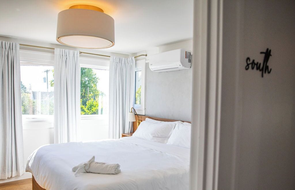 A hotel room with a large bed, white curtains, a ceiling lamp, an air conditioning unit, and a wall sign that says "south".