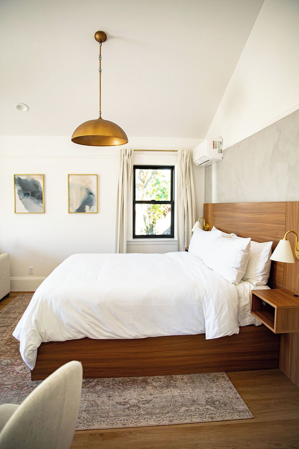 A bedroom with a large bed, a wooden headboard, a bedside table with a lamp, a hanging light fixture, and two abstract paintings on the wall.