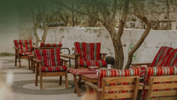 Outdoor seating with patterned cushions and wooden furniture, arranged under trees with a white wall in the background. - OliveTreeHostel (Ramallah)
