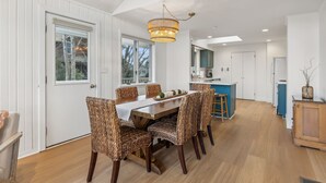 A dining area with a wooden table and woven chairs, a chandelier, and a view of the outdoors through large windows. - Walk to Kennebunk beaches! 4BR home with marsh views, large deck, and firepit (Kennebunk)