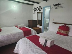 Habitación de hotel con dos camas, cada una con una manta roja y un almohadón blanco, un aparador de madera, un espejo y una puerta que da a un baño.