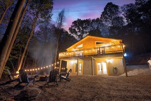 A wooden cabin with a balcony, surrounded by trees, and equipped with outdoor seating and string lights.