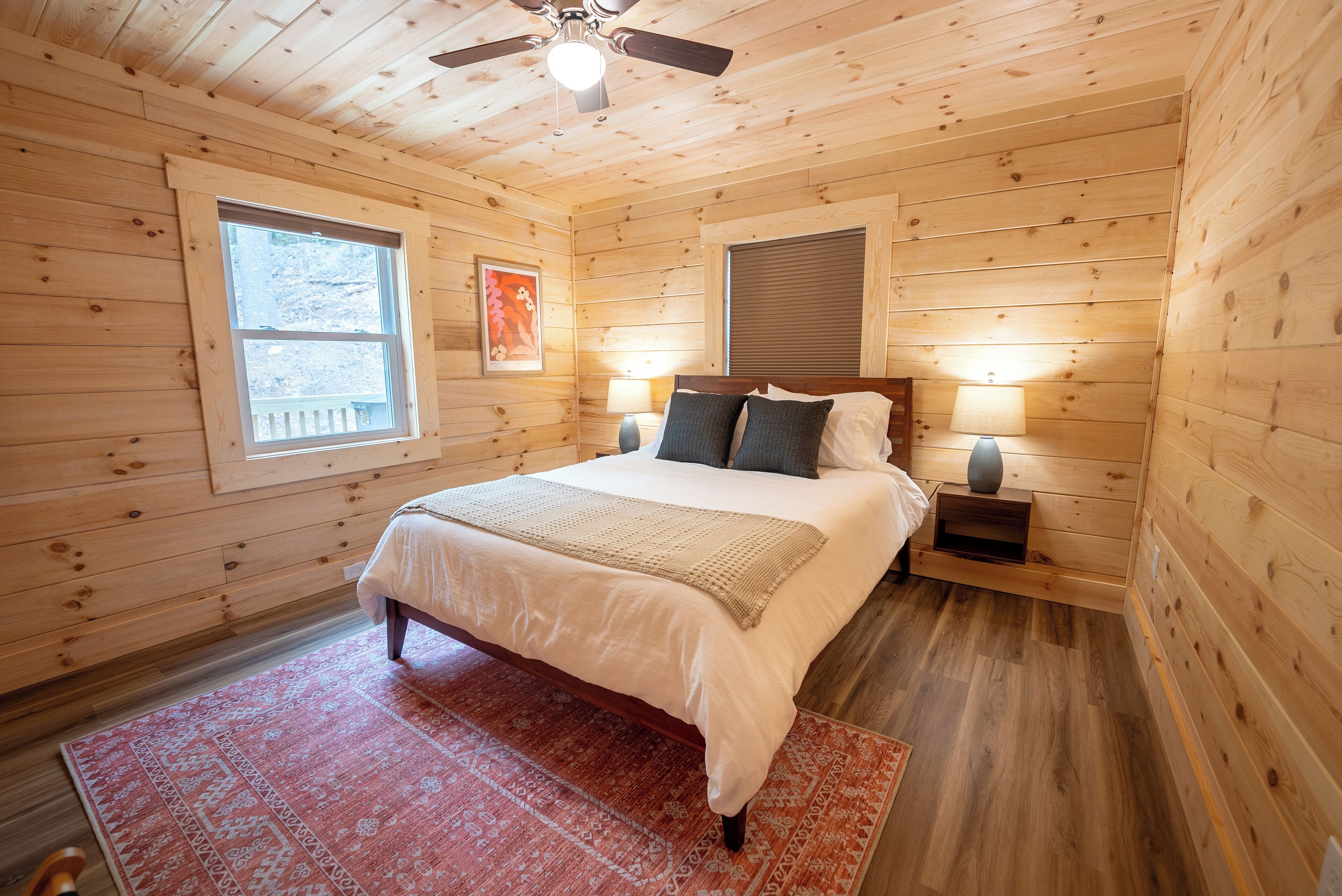 A wooden cabin bedroom with a bed, nightstands, a ceiling fan, and a window.