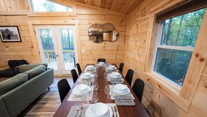 A wooden cabin dining room with a long table set for six, a green sofa, and a large window overlooking a forest.