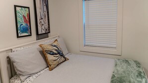 Une chambre à coucher avec un lit, deux oreillers, une fenêtre avec des stores et deux œuvres d’art encadrées accrochées au mur.