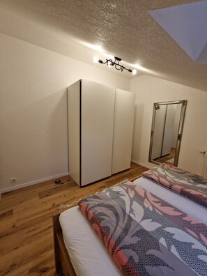 A bedroom with a wardrobe, a bed with patterned bedding, a ceiling light, and a mirror. - New living (Offenburg)