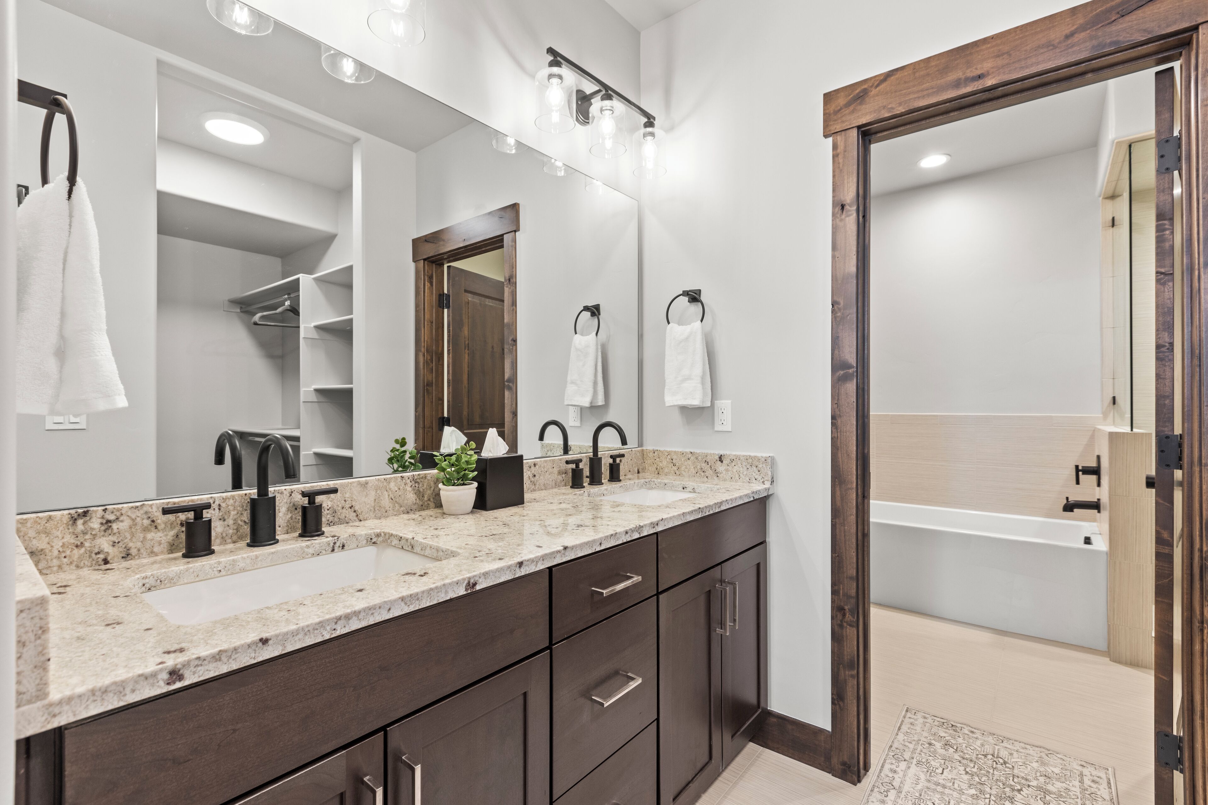 A modern bathroom with a double sink vanity, dark wood cabinets, a large mirror, and a wooden door leading to a shower area.