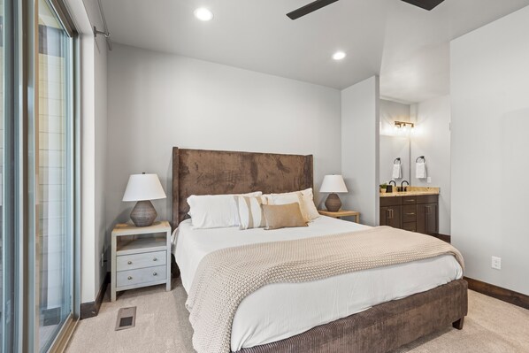 A bedroom with a large bed, two bedside tables with lamps, a wooden headboard, a bathroom with a sink and mirror, and a sliding glass door. - Black Rock Retreat Spacious 4-Bedroom Townhome (Kamas)