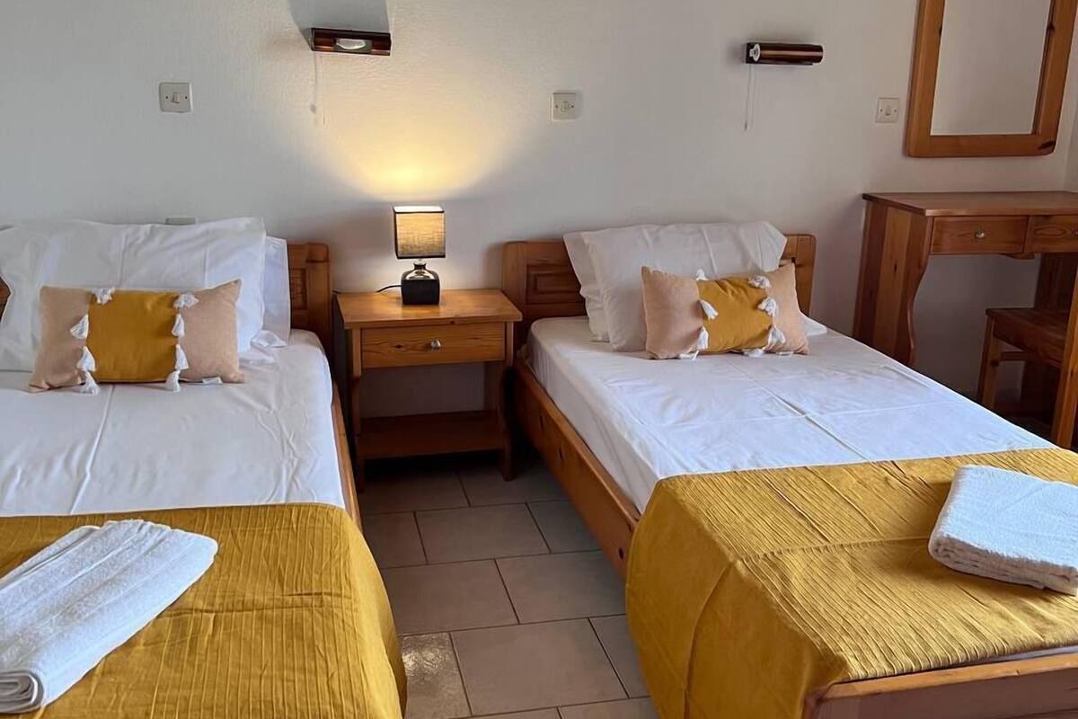 A hotel room with two single beds, each with a wooden headboard and a yellow bedspread. There is a nightstand with a lamp between the beds, and a wooden desk with a mirror to the right.