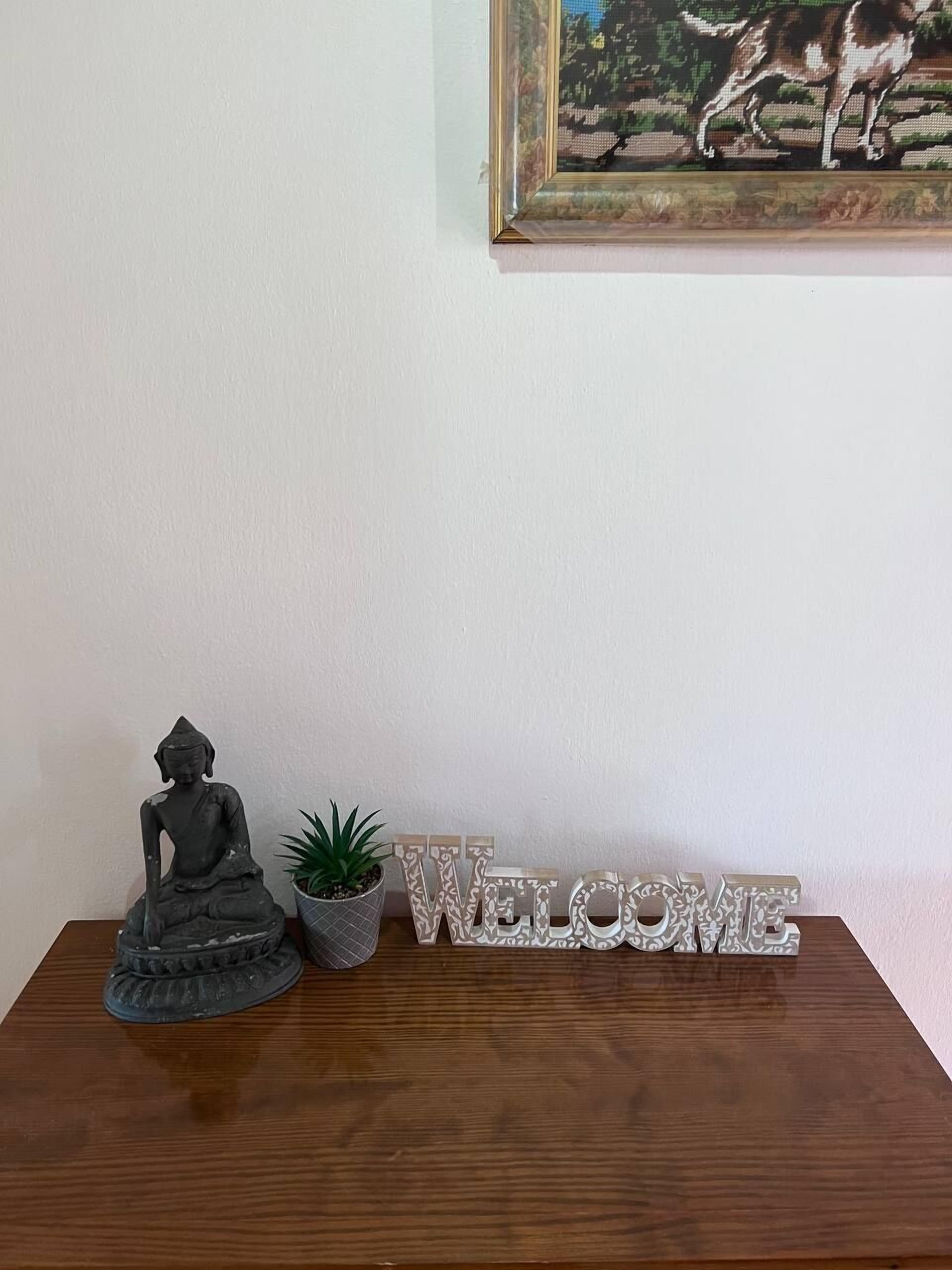 A statue of a seated figure, a small potted plant, and a 'WELCOME' sign on a wooden table against a white wall.