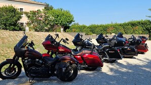 Una fila de motocicletas estacionadas sobre un terreno de grava, con un edificio y vegetación al fondo.