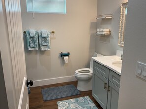 A bathroom with a toilet, sink, and shower. There are towels hanging on a rack and a small shelf with a roll of toilet paper.