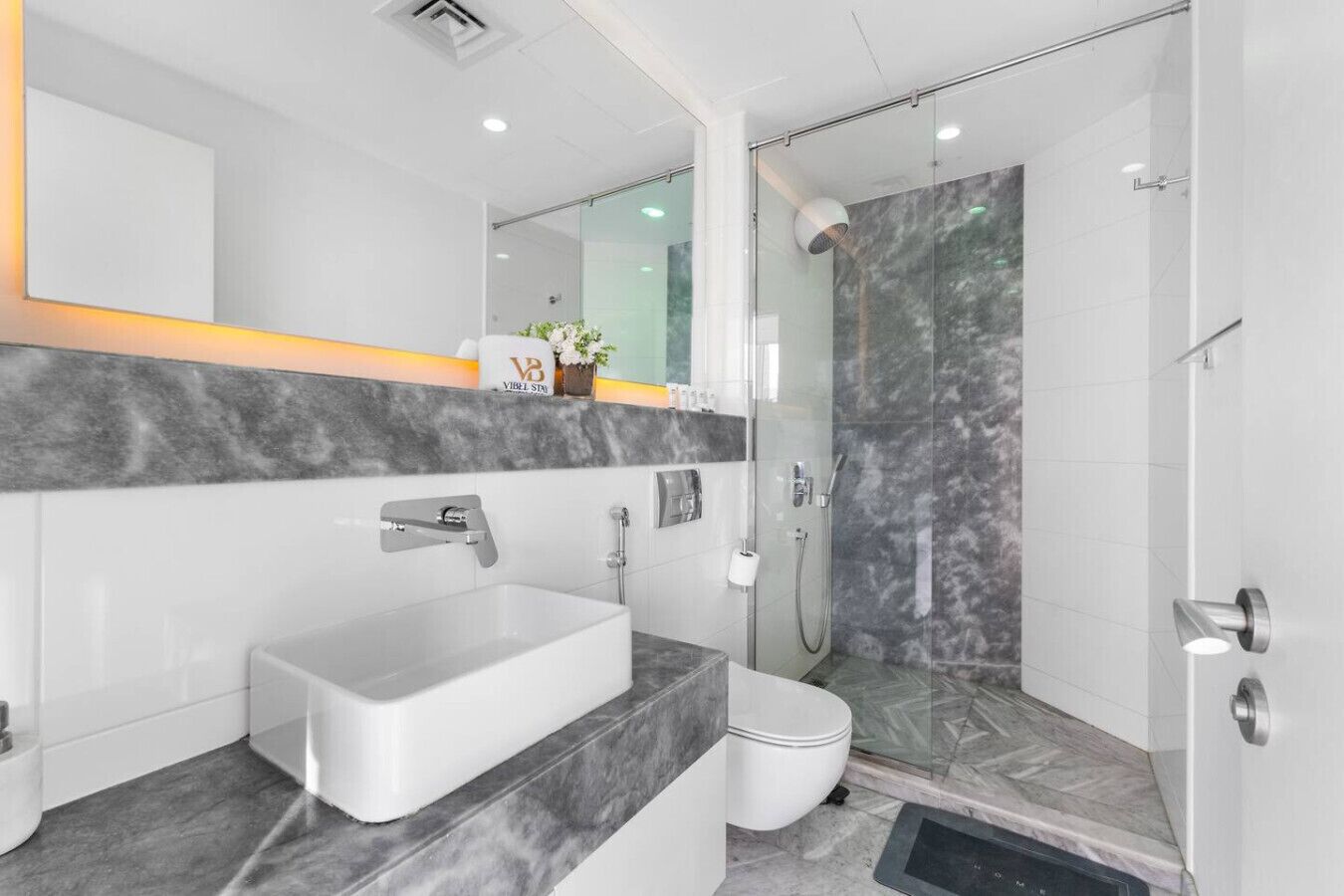A modern bathroom with a large mirror, a white sink, a shower, and a marble countertop.