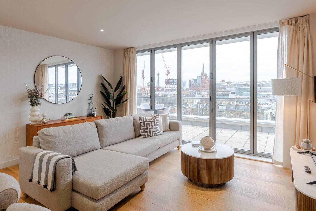 Ein modernes Wohnzimmer mit großem Fenster und Blick auf die Stadt, einem runden Holztisch und einem beige farbenen Sofa.