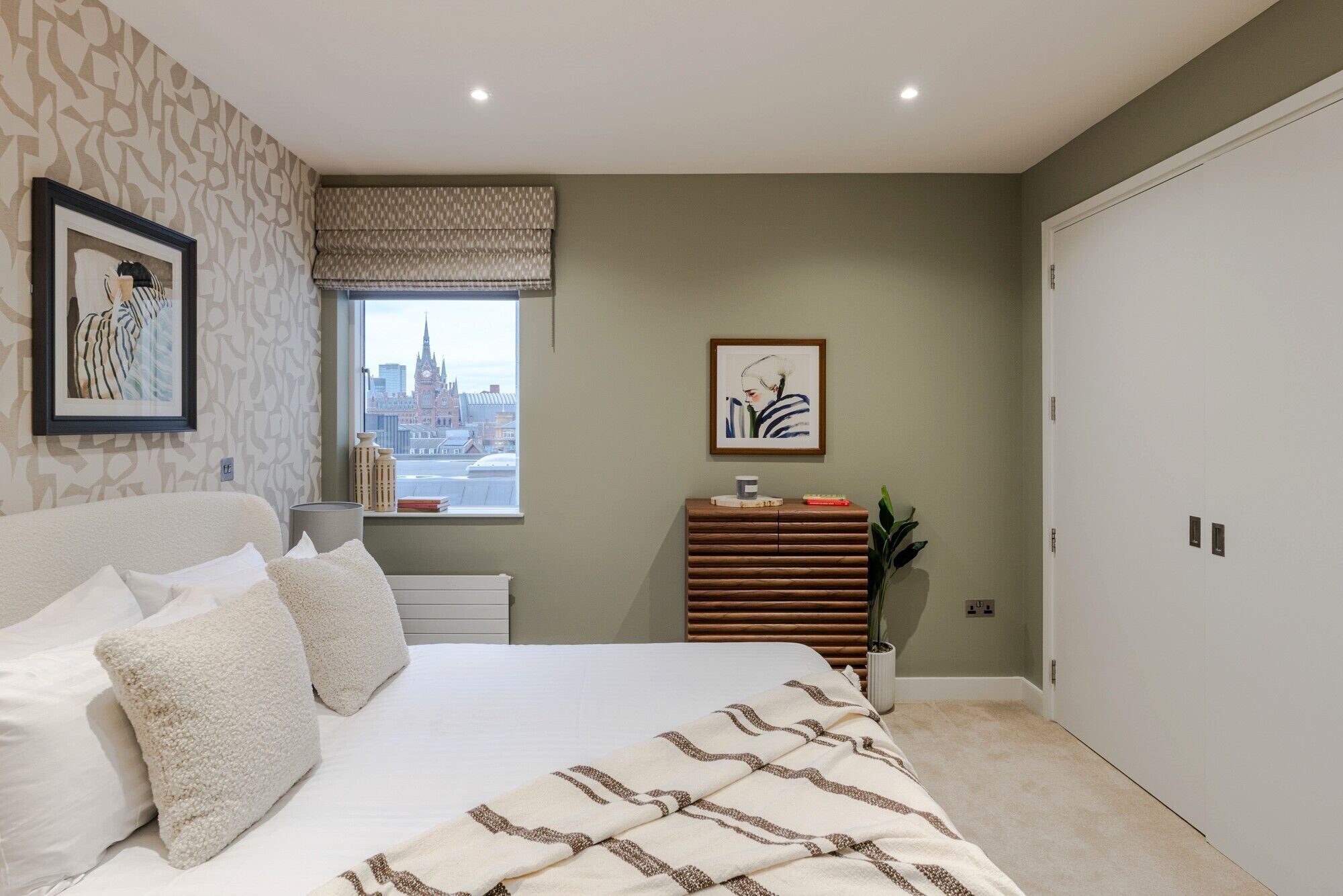 A bedroom with a bed, a window with a city view, a bedside table with books, and a wall-mounted artwork.
