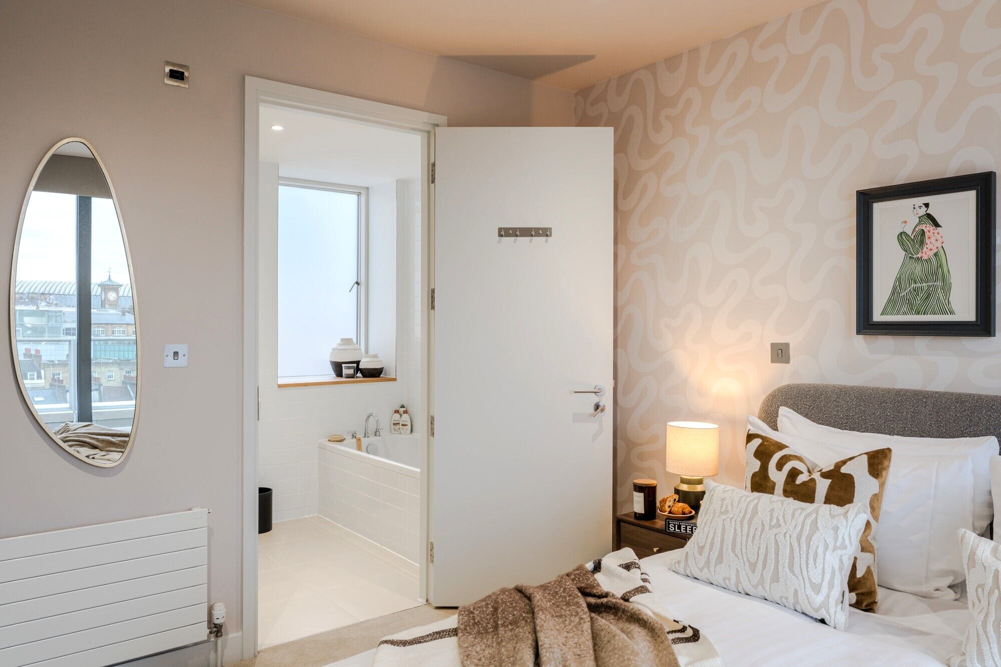 A hotel room with a large bed, a bedside table with a lamp, a framed picture on the wall, and a view of the city through a window.