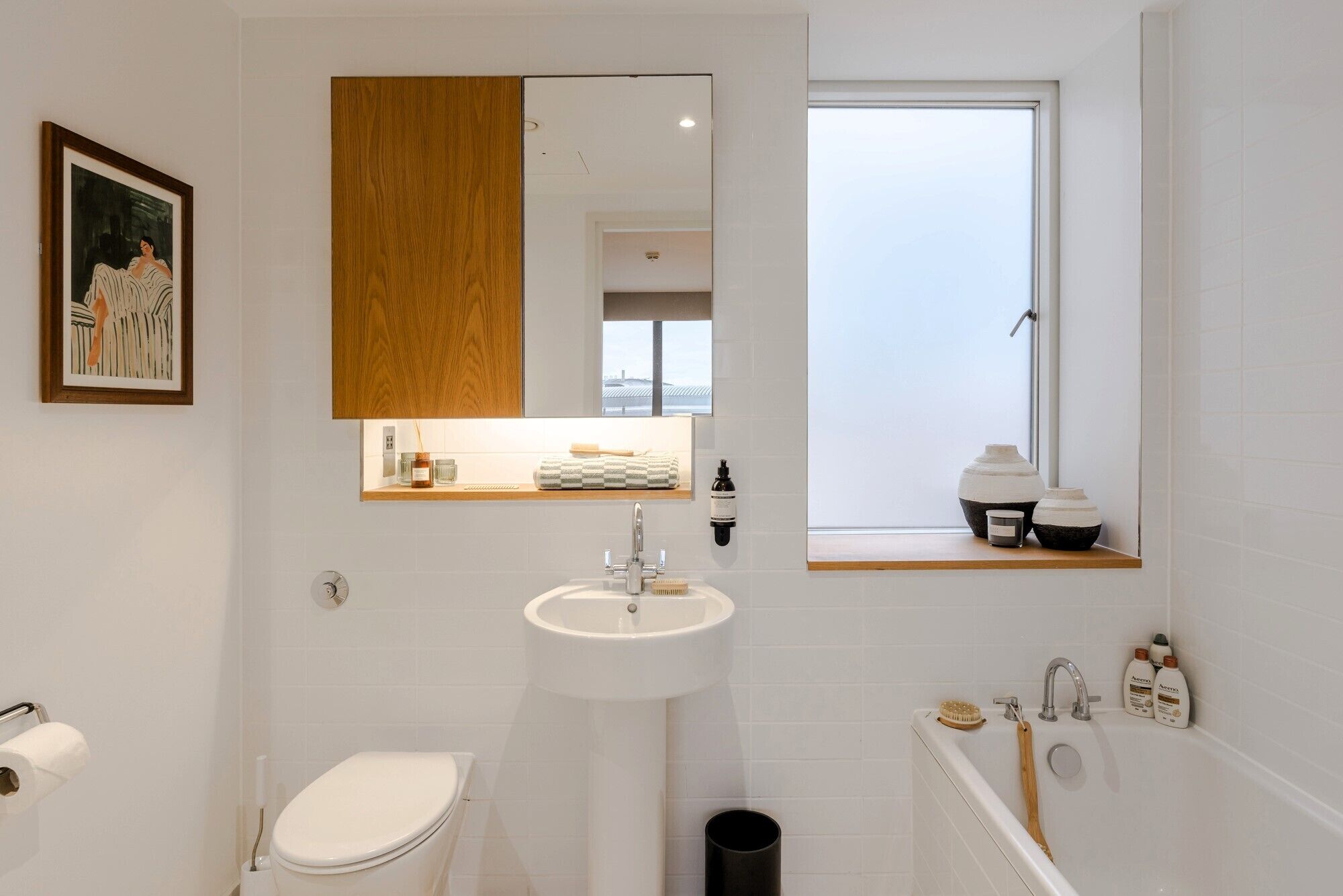Un baño moderno con inodoro, lavamanos y bañera. Hay una ventana sobre el lavamanos y un espejo sobre el inodoro.