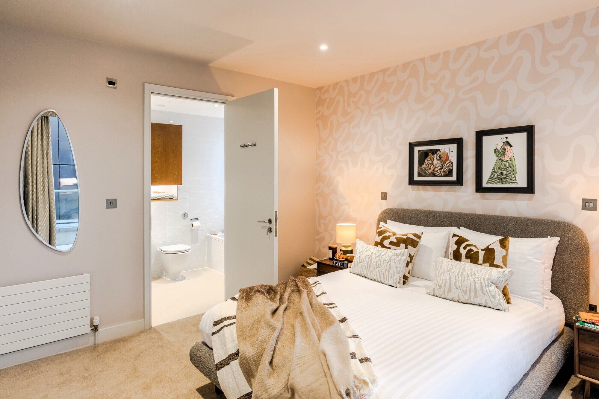 A bedroom with a bed, a bedside table with a lamp, a mirror, and a door leading to a bathroom.