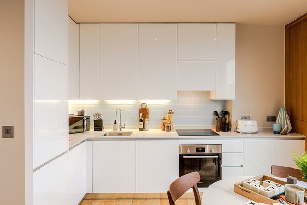 A modern kitchen with white cabinets, a built-in oven, and a dining area with a wooden table and chairs. - Your Apartment I King's Cross (London)