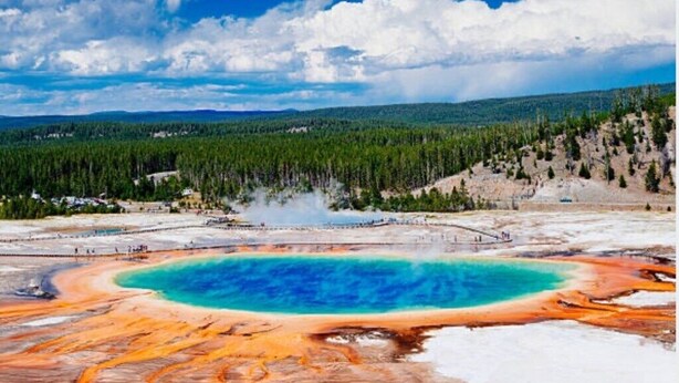 Un área geotérmica con una piscina turquesa, terreno circundante y colinas boscosas a lo lejos.