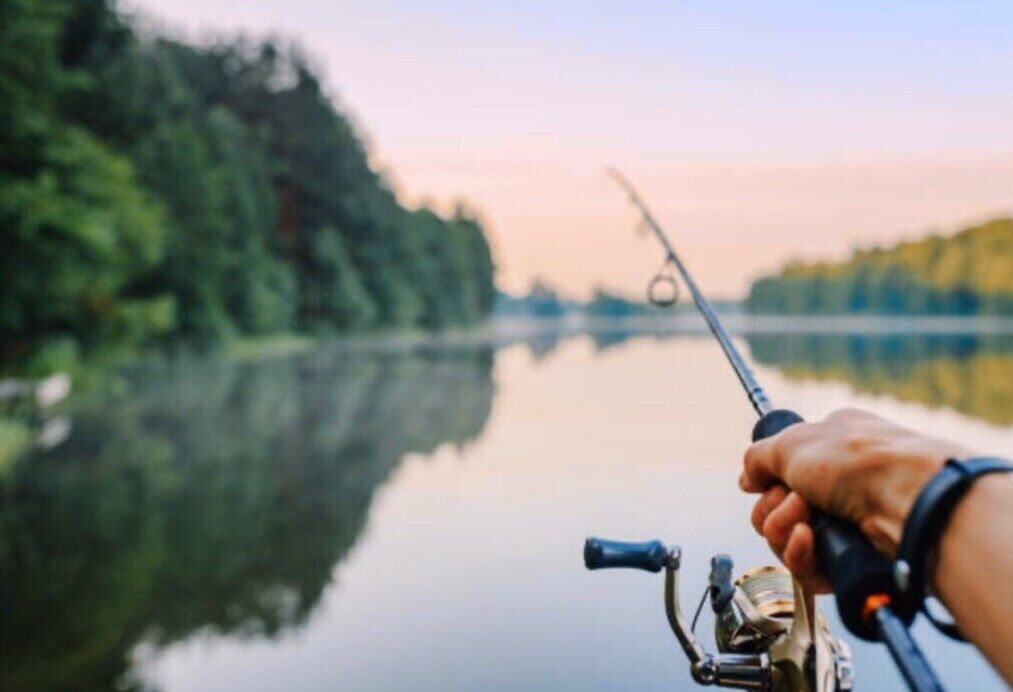 A hand holding a fishing rod over calm water with trees in the background.
