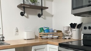 Eine Küche mit weißen Schränken, einer Mikrowelle, einem Herd und einer Kaffeemaschine. Über der Arbeitsplatte befinden sich Regale mit Glasbehältern und einer Pflanze.
