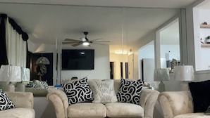 Ein Wohnzimmer mit einem beigefarbenen Sofagarnitur, gemusterten Zierkissen, einem Deckenventilator und einer modernen Kronleuchter.