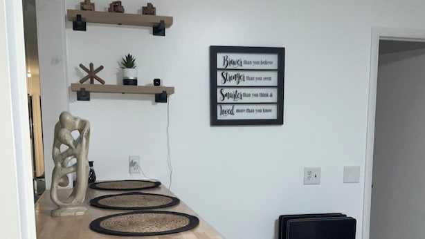 Ein Essbereich mit einem Holztisch, vier gewebten Tischsets, zwei dunklen Holzstühlen und einem schwarzen Rollstuhl. An der Wand über dem Tisch ist ein motivierendes Zitat angebracht.