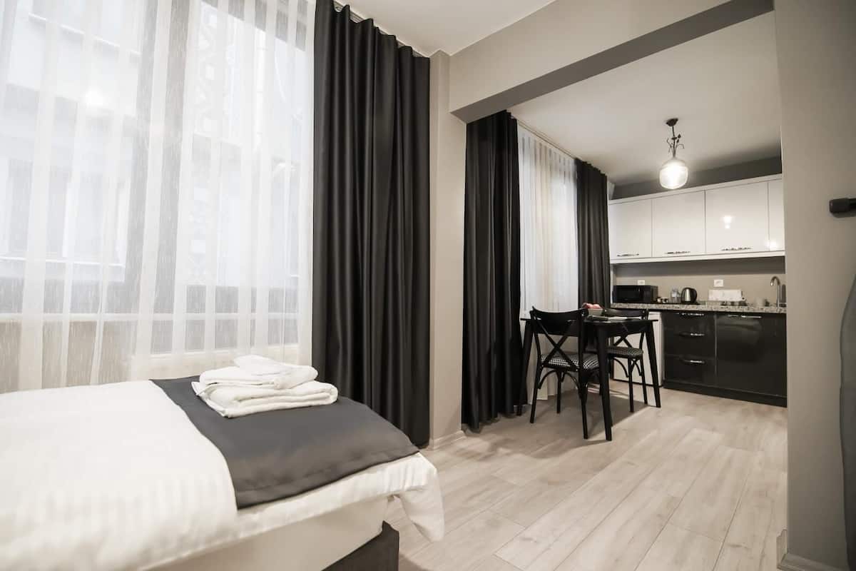 Una habitación de hotel moderna con cama, una mesa de comedor con sillas, zona de cocina y ventanas amplias con cortinas translúcidas.