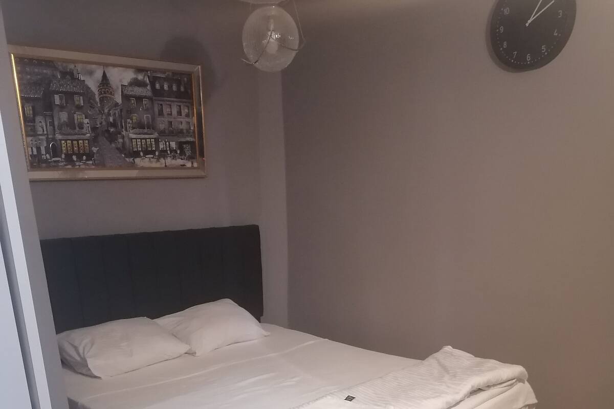 A neatly made bed in a room with a painting on the wall, a clock, and a ceiling light.