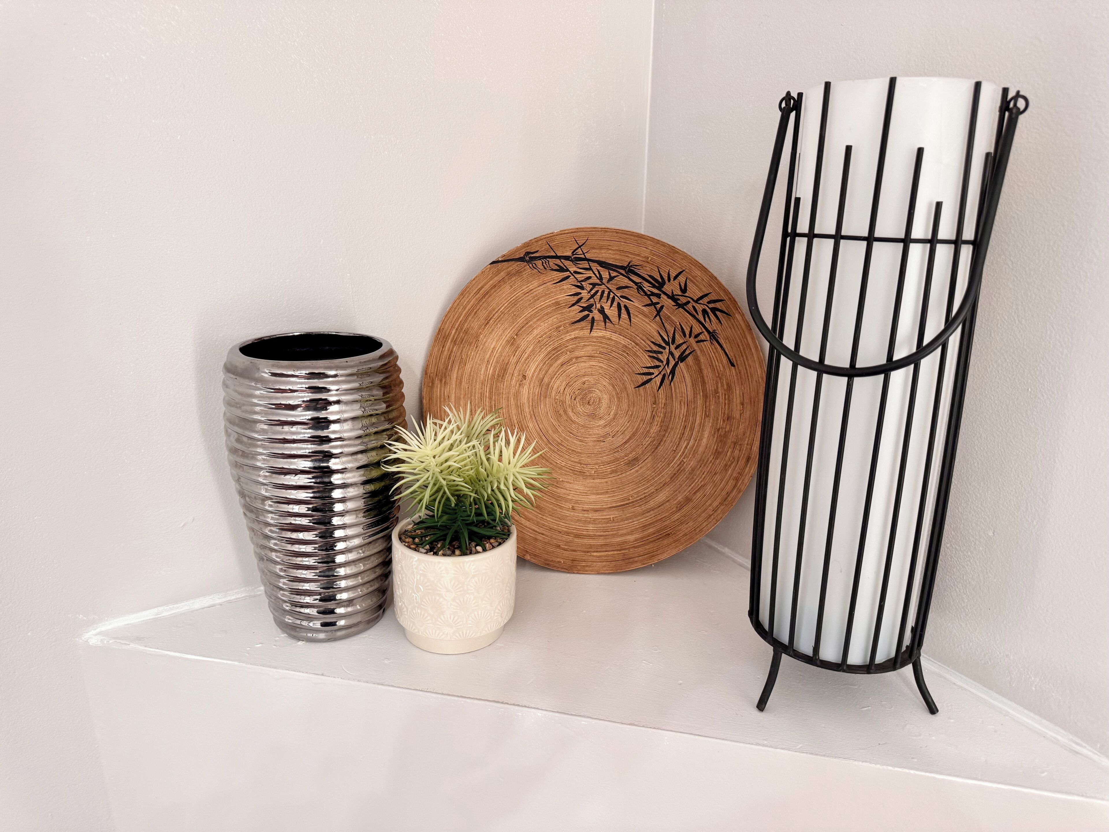 Un coin avec une étagère blanche où sont disposés un vase métallique, une petite plante en pot, un plateau rond en bois orné de motifs en bambou et un présentoir métallique noir.