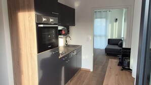 A modern kitchen with dark cabinets, a built-in oven, and a microwave. A dining table with chairs is in the foreground, and a sofa is visible in the background.
