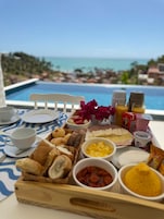 Bandeja de desayuno con diversos alimentos y condimentos junto a una piscina con vista al océano.
